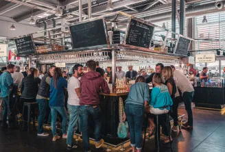 Interior view of Halles de Bacalan in Bordeaux showing a crowd of visitors gathered around a wine bar. Suspended glass shelves and menu chalkboards are visible above the counter in this modern covered market.
