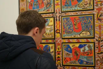 A child seen from behind wearing a black jacket gazes at a vibrant multi-panel artwork featuring geometric and figurative patterns in red, blue, yellow, and orange at the Musée de la Création Franche in Bordeaux.