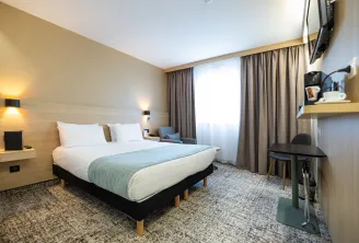 Modern hotel room featuring a double bed with white linens and light blue bedspread, light wood headboard, wall sconces, work desk with chair, gray curtains, and wall-mounted TV at Sure Hotel by Best Western Bordeaux Lac.