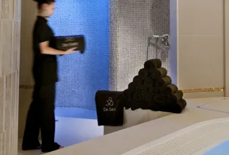 Interior of the Hôtel de Sèze spa in Bordeaux featuring a staff member in black attire near a blue water basin, stacked black towels, and textured tile panels in the background.
