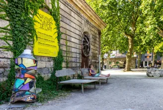 Stone courtyard with ivy-covered historic buildings, yellow « Become art supplies » poster, colorful sculpture, public benches and person sitting, trees in background