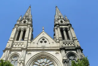 Front façade of Saint-Louis Church in Bordeaux featuring twin Gothic spires with pointed arches, a central rose window, and detailed stone architectural elements against a clear blue sky.
