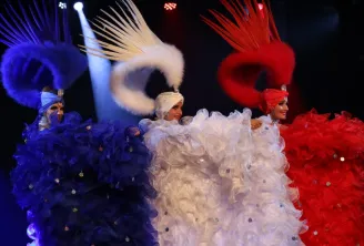 Three cabaret dancers on stage wearing elaborate blue, white, and red costumes with ornate headdresses and voluminous feathered backpieces, lit by colored spotlights at Le Grain d'Folie in Bordeaux.