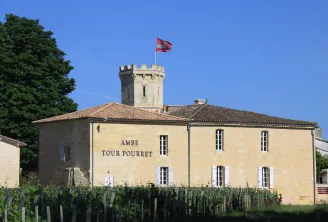 Ambe Tour Pourret castle in Bordeaux, a stone building with a crenellated tower topped by a French flag, surrounded by vineyards and trees against a blue sky.