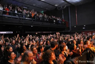 Enthusiastic crowd at a concert at Krakatoa in Mérignac, Bordeaux, with spectators applauding in the venue and fans in the upper seating area.