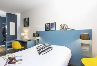 Modern bedroom at Ténéo Apparthotel Bordeaux Saint-Jean featuring a white bed, blue headboard, yellow chairs, integrated desk, and minimalist décor.
