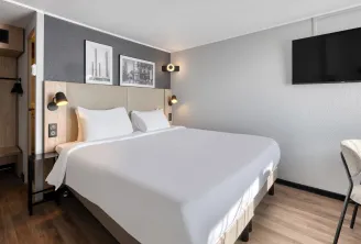 Modern hotel room featuring a double bed with beige headboard, black and gold wall lights, wall-mounted TV, minimalist grey and white décor, and a light upholstered chair