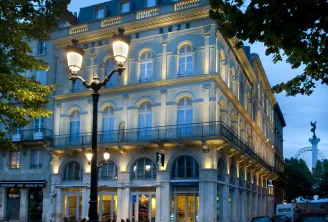 Illuminated facade of the Hôtel de Sèze in Bordeaux, a historic Haussmann-style building featuring columns, wrought-iron balconies and mansard roof, photographed at dusk with a street lamp in the foreground.