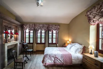 Elegant bedroom at Château Giscours featuring a bed with red and white checkered bedding, ornate fireplace, panoramic windows overlooking the vineyard, patterned curtains and classic furniture