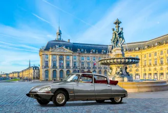 A vintage grey Citroën DS with red interior parked at Place de la Bourse in Bordeaux, featuring the Three Graces fountain and historic buildings in the background