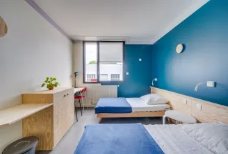 Double room at Bordeaux Youth Hostel featuring two single beds, turquoise and white walls, light wood furniture, city view window, small desk and modern amenities.