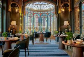 Elegant restaurant dining room featuring ornamental glass windows, round wooden tables, blue chairs, golden wall lamps, and a geometric glass dome ceiling with colorful panes.