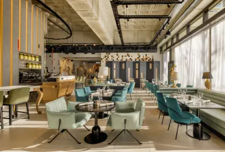 Modern and bright interior of Bada restaurant in Bordeaux featuring turquoise and green seating, black tables, wooden bar, large windows, industrial ceiling and gold geometric decor.