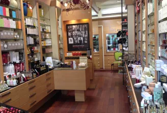 Elegant interior of a perfume shop featuring a central counter, light wood shelves displaying fragrance bottles, decorative chandelier, and artist photographs on the wall.
