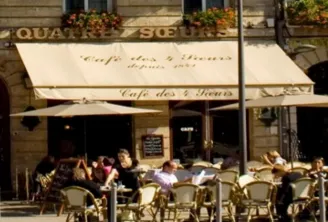 Sunny terrace of Café des 2 Fleurs in front of the Hôtel des Quatre Sœurs in Bordeaux, with customers seated at tables and historic stone façade.