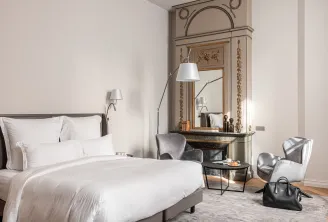 Elegant bedroom at Villas Foch hotel in Bordeaux featuring a king-size bed with white linens, ornate mirror above fireplace, gray armchairs, modern arc lamp, and refined classic décor