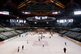 Interior view of Mériadeck Ice Rink in Bordeaux with multiple skaters on the ice, empty stands, and metal roof structure with orange and yellow lighting.