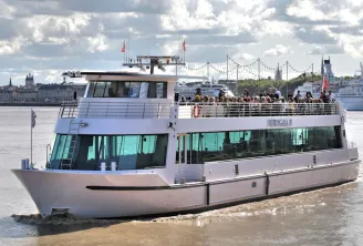 The Burdigala II cruise ship sailing on the Garonne River in Bordeaux, with passengers on the upper deck and the historic city skyline in the background.