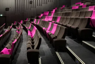 Modern cinema interior with black and hot pink recliners arranged in rows, pink architectural lighting on armrests, grey striped walls, and visible stairs in the background at Blanquefort, Bordeaux.