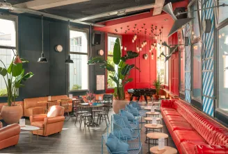 Modern lounge space in Bordeaux featuring red and teal walls, orange and red armchairs, blue chairs, gold hanging lights, green plants, and contemporary furniture in a vibrant urban setting.