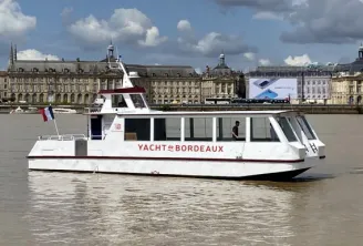 The Yacht of Bordeaux, a white cruise boat sailing on the Garonne River with 18th-century Bordeaux architecture in the background under a cloudy sky.