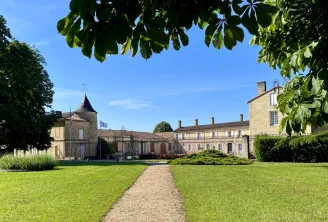 Courtyard of Château de Portets near Bordeaux featuring stone buildings with a conical tower, central pathway, and manicured green lawns against a clear blue sky.