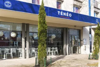 Facade of Ténéo Aparthotel in Talence Centre, Bordeaux, featuring modern large windows, blue and white signage, outdoor terrace with white chairs and green topiary plants.