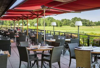 Covered terrace of Le Club restaurant at Golf du Pian Médoc in Bordeaux, featuring red umbrellas, wooden tables, black wicker chairs, and views over the lush fairways of the golf course.