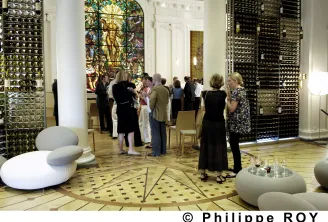Interior of the CIVB Wine Bar in Bordeaux showing visitors standing in conversation, surrounded by metallic display panels, a colorful stained glass window, and white rounded seating sculptures in the foreground.