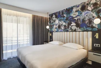 Modern hotel room featuring a double bed with white linens, beige upholstered headboard, black accent wall with blue and white floral mural, brown curtains, window with sheer drapes and round pendant lights.