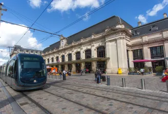 A modern blue tram approaches Bordeaux's Saint-Jean station, with the grand historic cream-colored building featuring ornate architecture and slate roof in the background under a clear blue sky.
