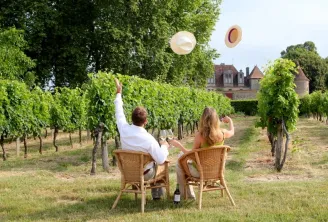 Two people sitting in wicker chairs in a vineyard, tossing their hats in the air. A historic castle is visible in the background.