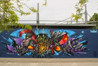 Colorful street art mural on the M.U.R wall in Bordeaux, featuring stylized faces with geometric shapes in blue, red, gold, and purple tones on a dark blue background, beneath a modern bridge.