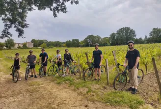 A group of cyclists with their gravel bikes posing on a dirt road between Bordeaux vineyards, with rows of green vines and stone buildings in the background.