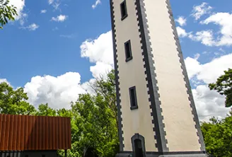 Tall white and black tower on Patiras Island in Bordeaux, surrounded by green lawn and a red building in the background, under a partly cloudy blue sky.