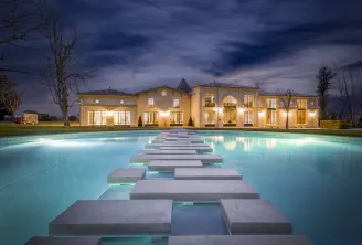Night view of Château Montlabert in Bordeaux with its golden-lit façade, adjacent to a turquoise pool crossed by a symmetrical gray stone pathway, beneath a dramatic twilight sky.