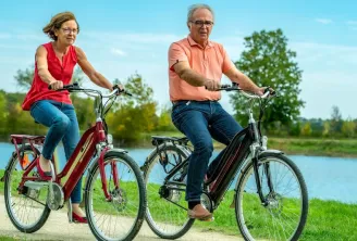 Senior couple riding electric bikes on a path near water in Bordeaux, sunny day with trees in the background
