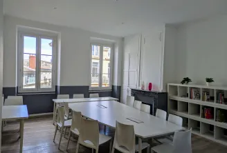 Modern workspace at FL Studios in Bordeaux featuring white tables, beige chairs, built-in shelving, black fireplace, and large windows overlooking the city.