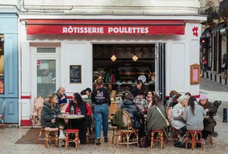 Exterior view of Rôtisserie Poulettes in Bordeaux with a bustling outdoor terrace featuring customers seated at bistro tables enjoying meals in front of the red and white striped storefront.