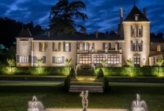 Château Bellefont Belcier in Bordeaux lit up at night, featuring a pale stone facade with pointed towers, central access stairs and illuminated fountains in the foreground