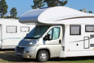 Three white motorhomes parked side by side in a parking area, with coniferous trees in the background and a clear blue sky.