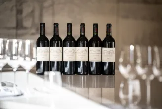 Six bottles of red wine from Château de Ferrand vintage years 2012 to 2017 lined up on a white table with blurred wine glasses in the background, in an elegant Bordeaux setting.