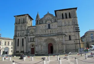 The Abbey Church of Holy Cross in Bordeaux, a Romanesque church with two square towers, ornate stone façade and rounded main portal, located on a paved square with safety barriers, under clear blue sky.