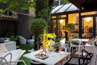 Elegant terrace of a boutique hotel in Bordeaux featuring white dining tables, modern chairs, landscaped garden, ambient lighting, and illuminated glass doors opening to the restaurant-bar interior.