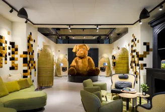 Modern lobby of FirstName JDV by Hyatt hotel in Bordeaux featuring a large teddy bear seated in the center, surrounded by green armchairs, rattan floor lamps, and walls decorated with yellow and black tiles.