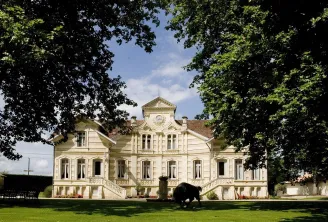 Front view of Château Maucaillou, a historic Belle Époque-style building with white façades and balusters, surrounded by lush trees and a manicured lawn. A black horse grazes in the park.