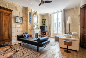 Elegant living room in a Haussmann apartment in Bordeaux featuring exposed stone walls, gilt fireplace, contemporary black and beige furniture, large windows overlooking the street, and refined decor.