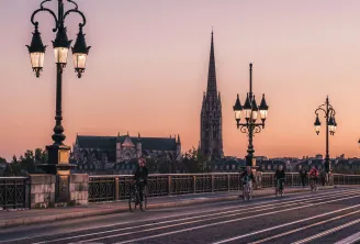 Stone Bridge in Bordeaux at sunset with Saint-Michel Spire in the background, cyclists crossing the illuminated bridge with ornamental street lamps.