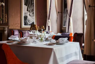 Elegant fine dining restaurant room with round table setting, burgundy and orange upholstered chairs, fine glassware, white linens and sophisticated drapes at Le Pressoir d'Argent Gordon Ramsay in Bordeaux.