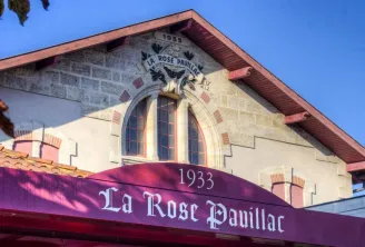Front facade of La Rose Pauillac wine cellar in Bordeaux, built in 1933, featuring stone architecture, arched windows, and a purple storefront sign.